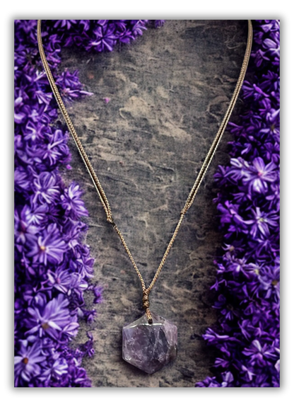 A gold-tone chain necklace with a raw, faceted purple crystal pendant, displayed on a textured gray stone surface and framed by purple flowers on both sides.