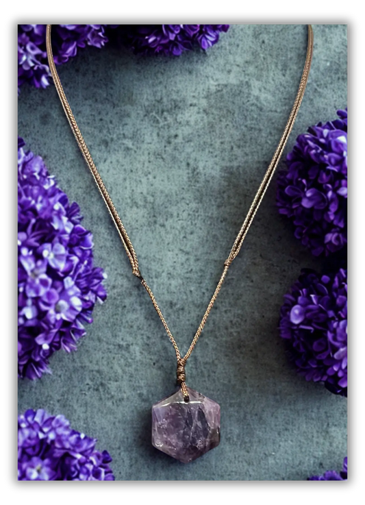 A rose gold chain necklace with a faceted purple crystal pendant, styled on a textured gray background with purple flowers.