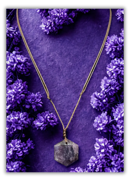 A gold chain necklace with a faceted gray crystal pendant, displayed on a textured purple background surrounded by small purple flowers.