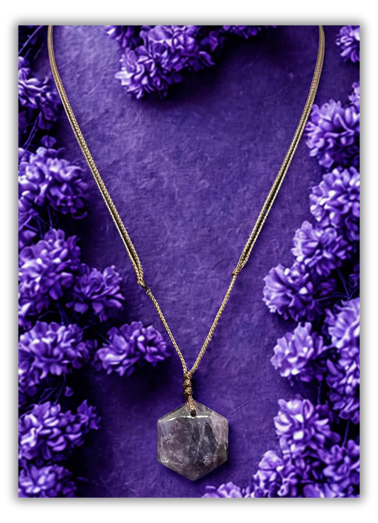 A gold chain necklace with a faceted gray crystal pendant, displayed on a textured purple background surrounded by small purple flowers.
